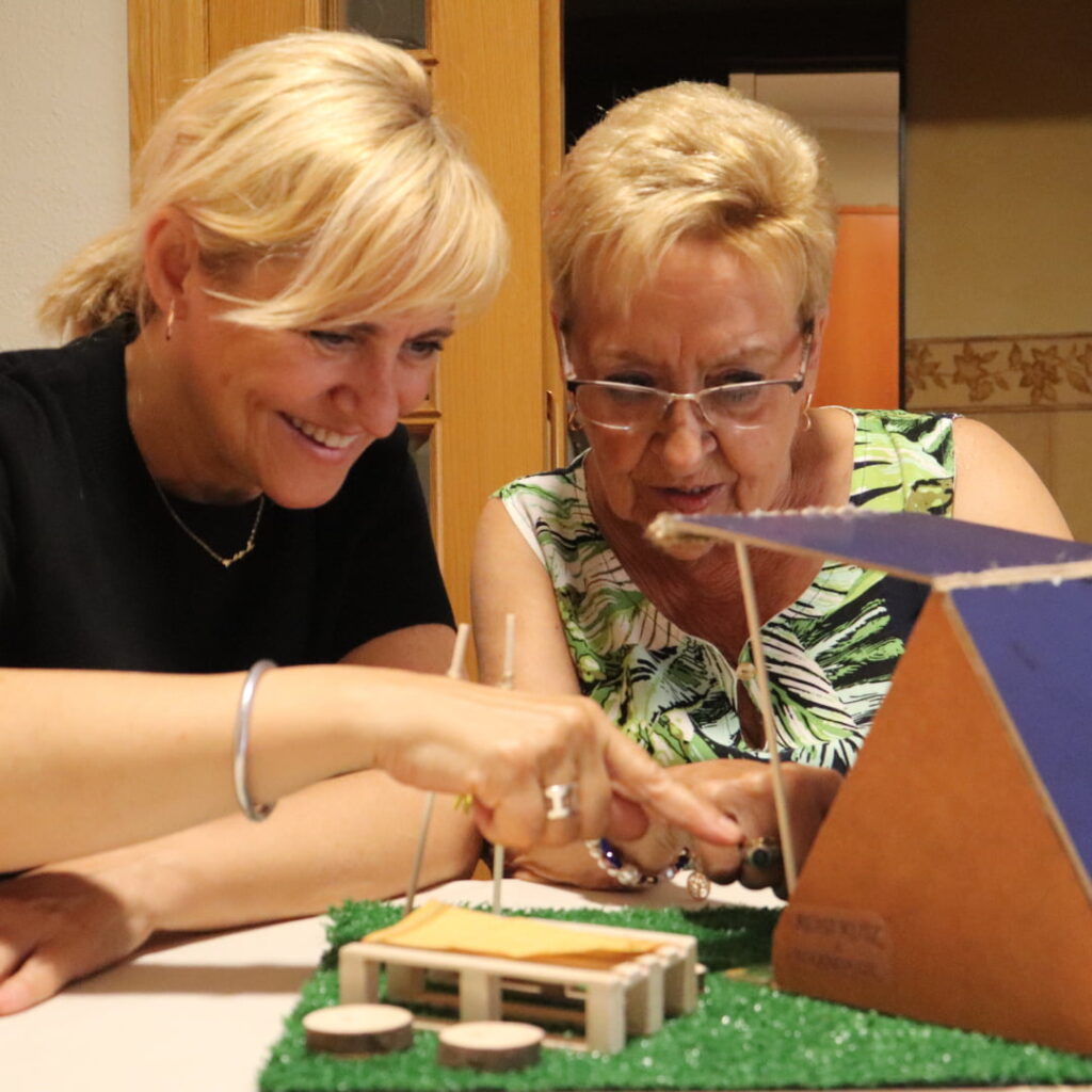 Imagen de dos mujeres, una mayor y una joven, jugando juntas a juegos de memoria en casa de la mujer mayor. La escena transmite cercanía, compañía y el fortalecimiento de la memoria y la conexión emocional.