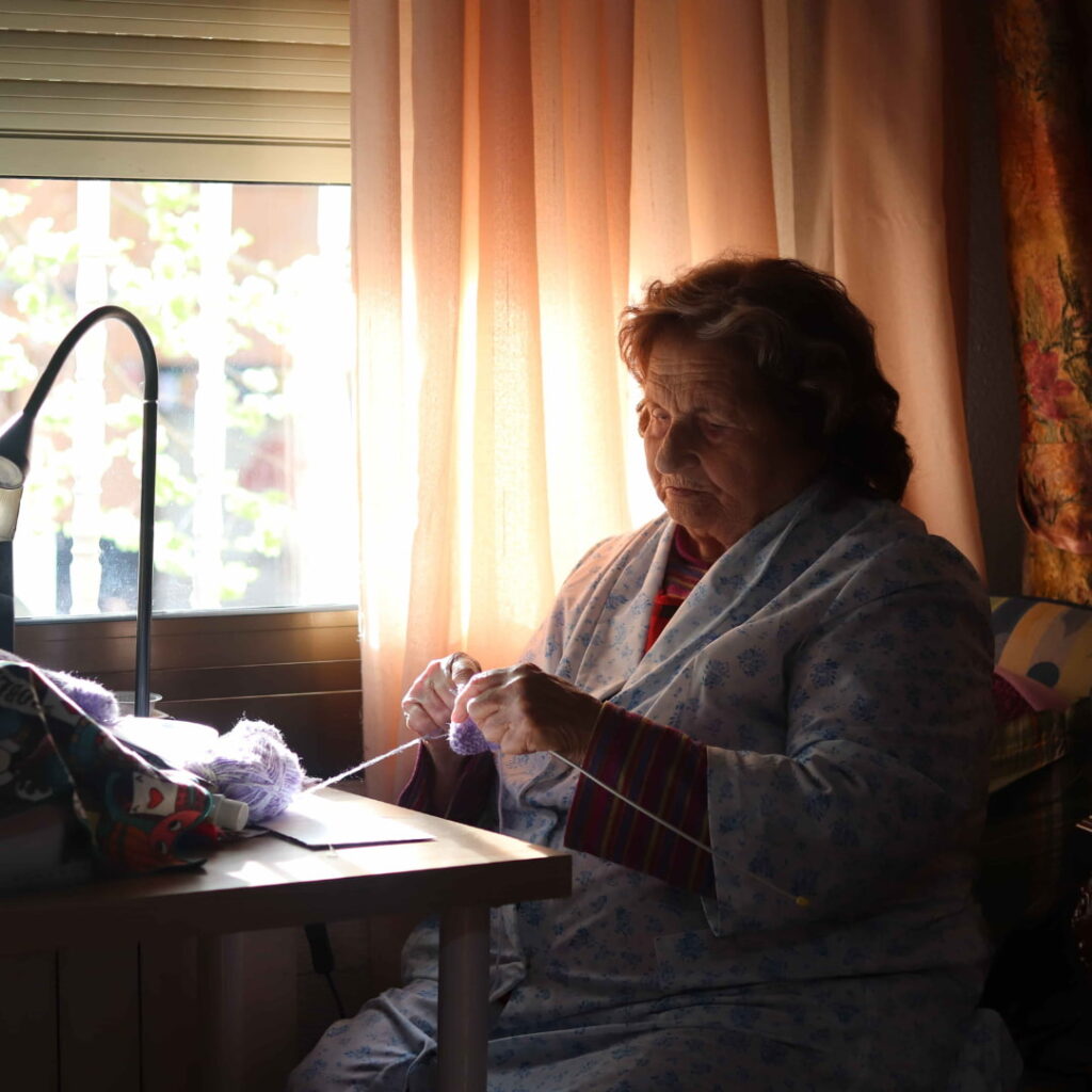 Imagen de una mujer mayor sentada en un rincón oscuro de su casa, tejiendo en soledad. La luz tenue de la ventana, a contraluz, resalta su tristeza, mostrando la soledad no deseada. Viste una bata de estar por casa, reflejando la quietud y el aislamiento que muchas personas mayores enfrentan en su día a día.